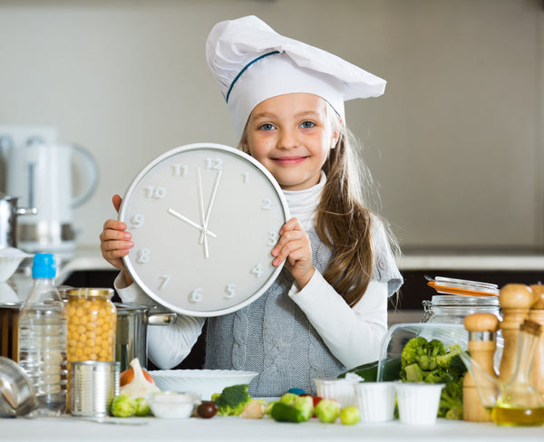 Girl,With,Ponytails,Holding,Clock,At,Kitchen,Table,And,Smiling